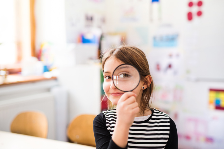 A small girl with a magnifying glass sitting at the desk at school.